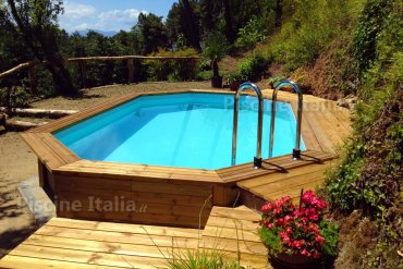 Piscina ottagonale allungata Jardin 5,60 m con rivestimento in legno e cornice chiara. Inserita in un contesto verde con siepi e prato, ideale per giardini residenziali.
